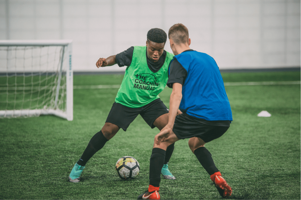 Two players contest a 1v1 in TCM bibs at an indoor pitch